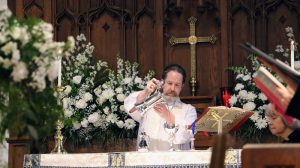 rector celebrating Eucharist on Easter surrounded by flowers. Cross mounted on oak-paneled church interior in the background.
